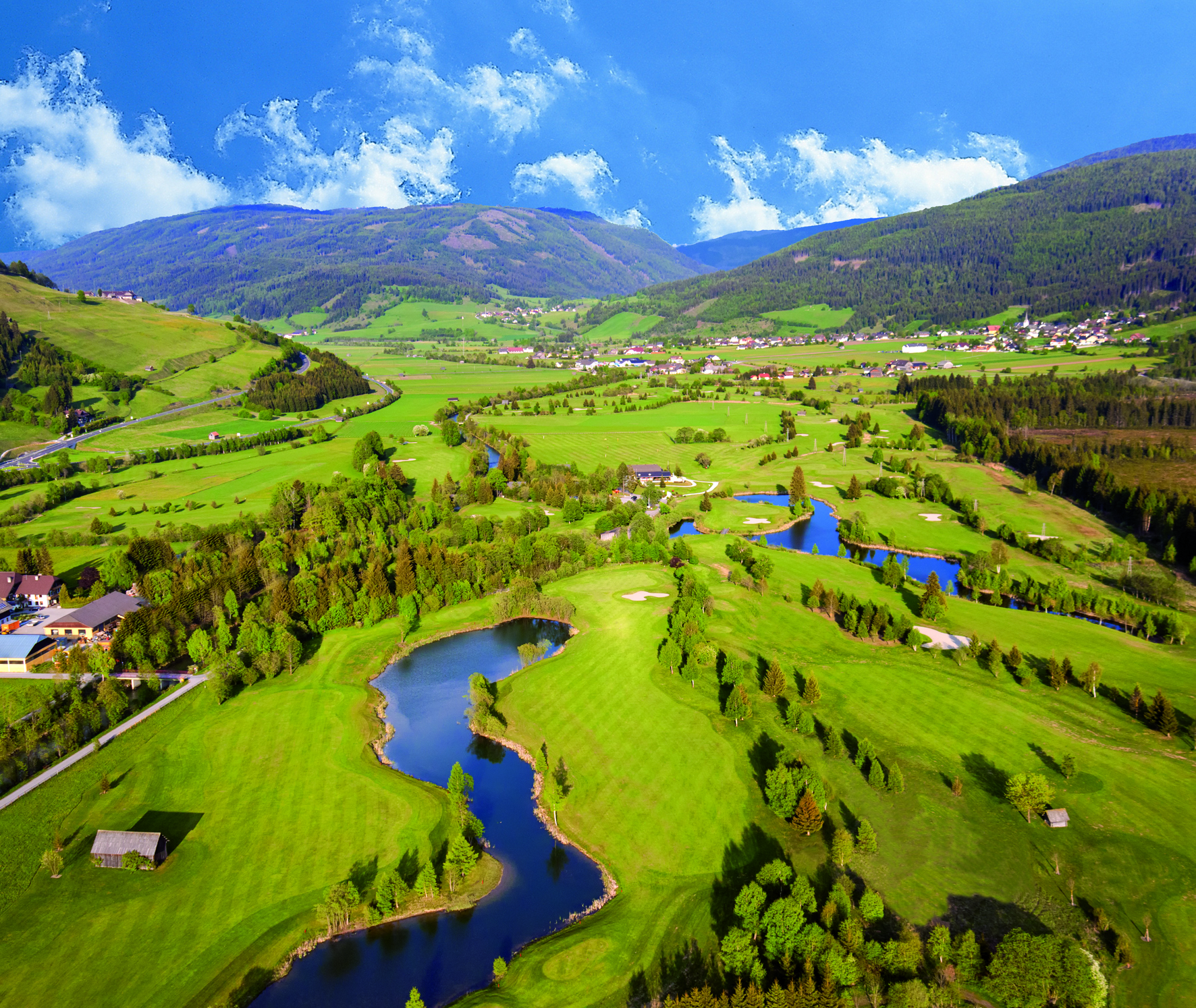 Luftaufnahme eines Golfplatzes mit grünen Wiesen, Bäumen und einem kleinen See, umgeben von Bergen und einem blauen Himmel.