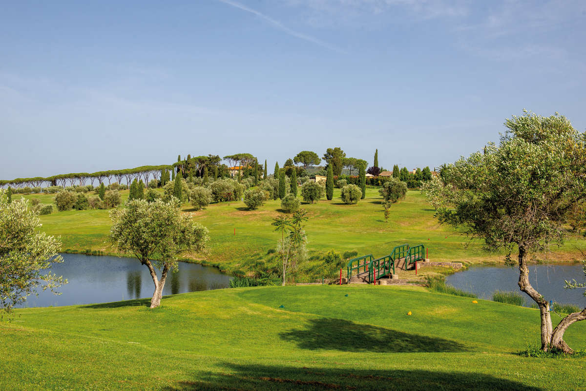 Landschaft mit grünem Golfplatz, zwei Seen und Bäumen, im Hintergrund Hügel und Zypressen.