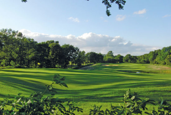 Weitläufige Golfplatzlandschaft mit grünem Rasen, Bäumen im Hintergrund und blauem Himmel.