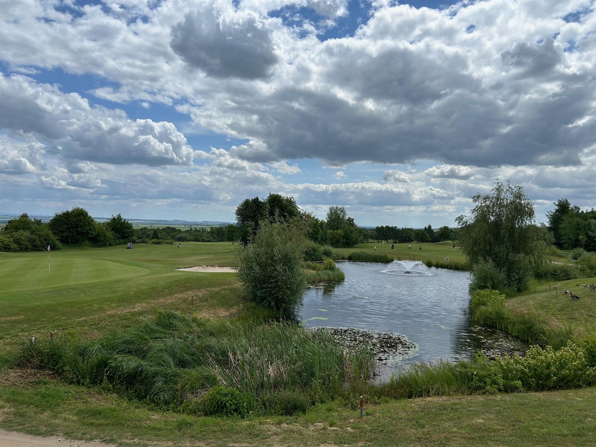 Golfplatz mit Teich, Springbrunnen und Sandbunker unter bewölktem Himmel.